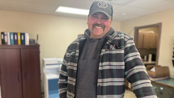 
Dans un bureau, un homme portant une grosse moustache, coiffé d'une casquette de baseball et vêtu d'une chemise à carreaux par-dessus un chandail à capuchon sourit à belles dents.