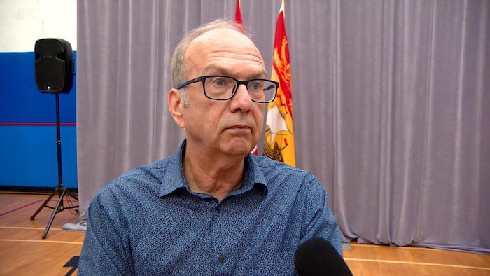 Un micro pointé en sa direction, un homme accorde une entrevue. Il est debout dans une salle, devant un rideau et un drapeau du Nouveau-Brunswick.