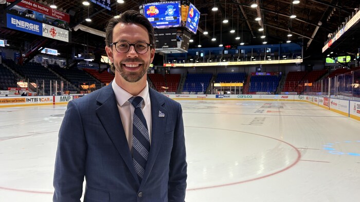 Un homme souriant à lunettes, vêtu d'un complet bleu et d'une cravate, pose directement sur la glace d'une patinoire. L'arrière-plan présente l'aréna vide avec ses estrades colorées, ses bannières publicitaires et son grand tableau indicateur suspendu.
