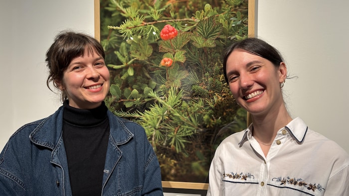 Les artistes Raphaëlle Ainsley-Vincent et Éloïse Lamarre sourient à la caméra. Elles se tiennent devant l'une des photographies de leur exposition représentant un grand plan sur une tourbière d'où émerge une chicoutai.