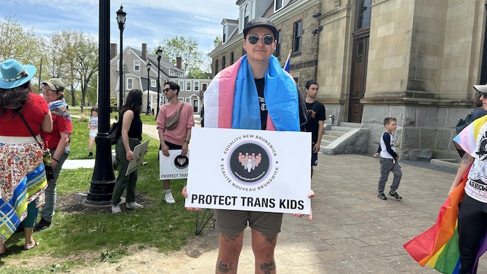 Une personne debout à l'extérieur, un drapeau autour des épaules, pose pour une photo en tenant une affiche sur laquelle est écrit « Protégez les enfants trans ».
