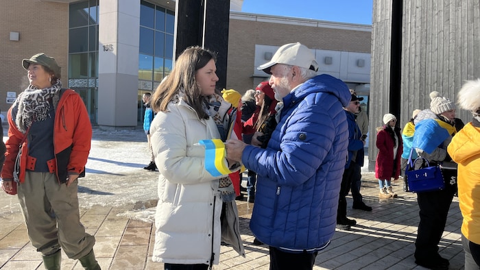 Une jeune femme en manteau blanc et un homme en veste bleue discutent lors d'un rassemblement extérieur ensoleillé. Plusieurs personnes à l'arrière-plan portent des accessoires aux couleurs du drapeau ukrainien, bleu et jaune, sur un sol pavé et enneigé.