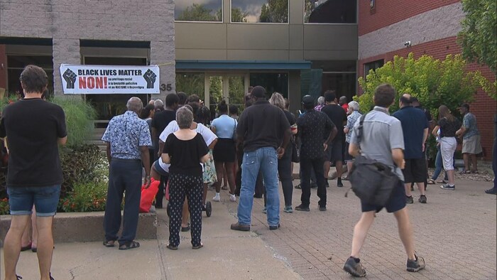 Des personnes rassemblées devant un édifice, tandis qu'on voit une bannière disant «Black Lives Matter, Non!» accrochée à un mur.