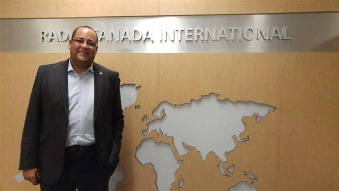 Saul Polo pose devant une carte du monde avec marqué "Radio Canada International" au dessus.