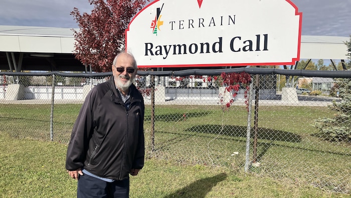 Raymond Call prend la pose en souriant près de la clôture et de l'affiche indiquant «Terrain Raymond Call».