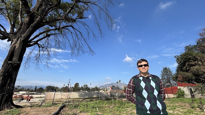 Raymond Francis pose devant un terrain d'où émergent des tournesols, un arbre et des restes d'habitations.