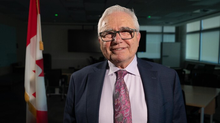 Raymond Théberge habillé d'un veston et d'une cravate se laisse prendre en photo devant un drapeau du Canada.