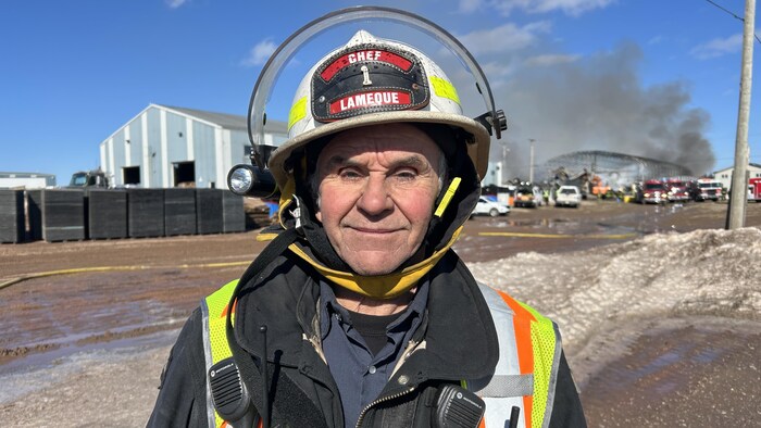Un pompier en uniforme sourit à la caméra, de la fumée et des camions de pompiers derrière lui.