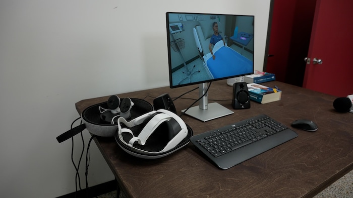 Un écran d'ordinateur montre un diligent  dans un lit. Il y a un clavier, un casque de réalité virtuelle et des manettes sur un bureau.