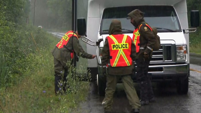 Trois policiers en habits imperméables dehors, sous la pluie, sur une route bordée d'arbres, à côté d'un petit autobus.