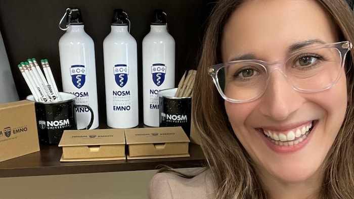 Portrait de Mélanie Goulet devant une étagère remplie de bouteilles, de tasses et de crayons avec le logo de l'École de médecine du Nord de l'Ontario.