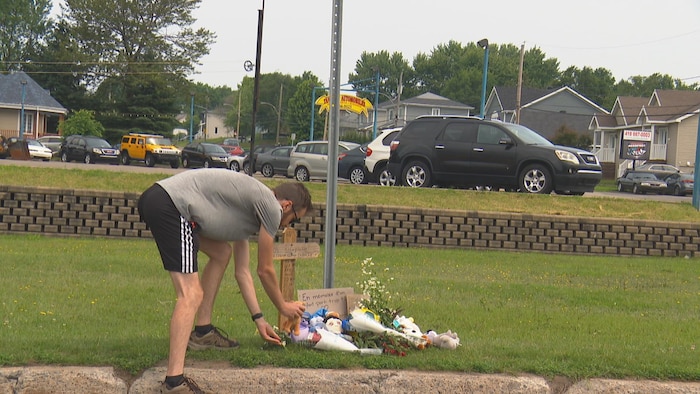 Un homme se penche pour déposer une peluche et des fleurs sur une pelouse.