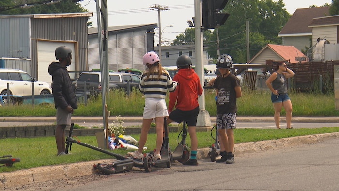 Plusieurs enfants en trottinettes électriques observent depuis la rue les gerbes de fleurs.