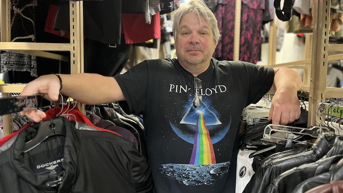 Un homme les bras posés sur des supports où sont accrochés des vêtements regarde l'objectif.