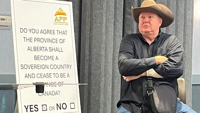 Jeff Rath est assis sur un tabouret à côté d'une affiche qui indique une question référendaire sur la souveraineté de l'Alberta.