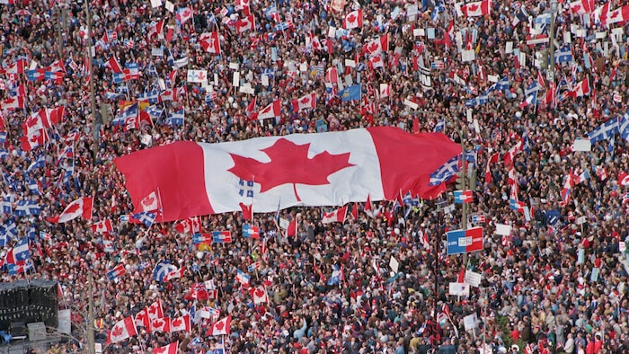Vue aérienne de milliers de personnes brandissant des drapeaux canadiens et québécois dont un drapeau géant du Canada