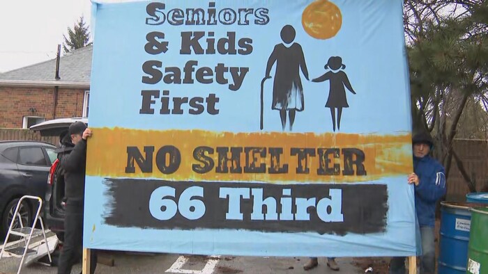 Deux hommes tiennent une banderole où il est écrit « Pas de refuge au 66 rue Third. La sécurité des aînés et des enfants doit passer avant.»