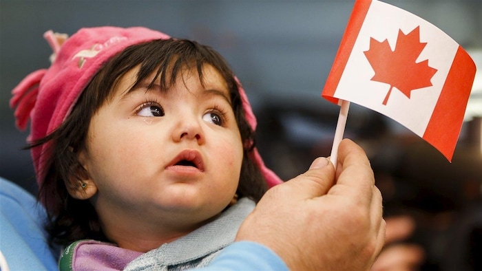 طفلة سورية تتأمل علم بلدها الجديد، كندا، في يد أبيها الذي يحملها عند وصول العائلة إلى مطار تورونتو بيرسون الدولي في 18 كانون الأول (ديسمبر) 2015 بعد حصول العائلة على حق اللجوء إلى كندا.