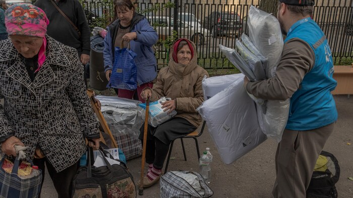 Une femme âgée est assise à un point d'évacuation et attend, entourée d'autres évacuées et d'un bénévole.