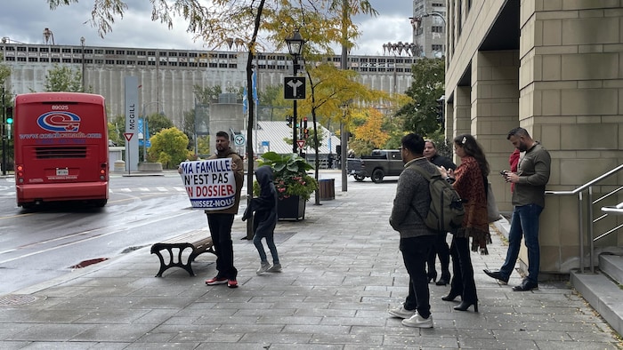 Sur le trottoir, un homme avec une affiche sur laquelle on lit « Ma famille n'est pas un dossier - Réunissez-nous ». Derrière lui, quelques individus