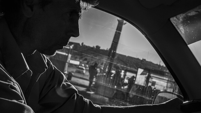 Un homme au volant d'une voiture et, au loin, des silhouettes d'hommes.