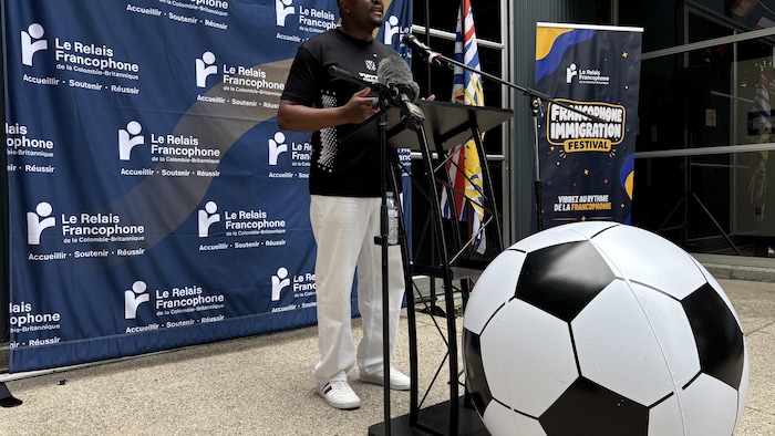 Franklin Bouguep s'exprimant devant un lutrin noir avec un microphone. Un ballon de soccer géant est posé au sol devant lui, sur fond de bannière bleue du Relais francophone. 
