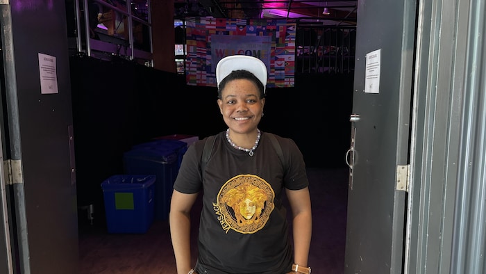 Lopez Mamoudou souriant de face, debout dans l'embrasure d'une porte double. Il porte une casquette blanche et un t-shirt noir orné d'un grand logo circulaire doré.