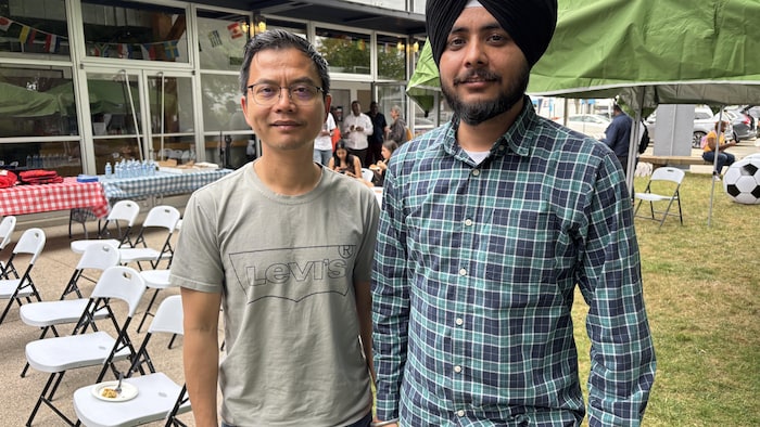 Deux hommes debout côte à côte à l'extérieur près d'un chapiteau. L'homme de droite porte un turban noir et une chemise à carreaux, et l'homme de gauche porte un t-shirt gris et des lunettes. 