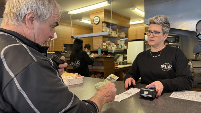 Un homme tend un billet de 20$ à une femme, derrière un comptoir.