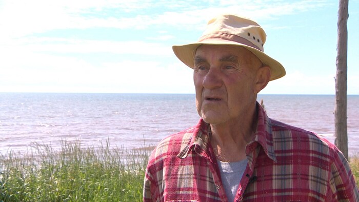 Un homme sur le bord de l'eau. Il est coiffé d'un chapeau mou et vêtu d'une chemise à carreaux rose.