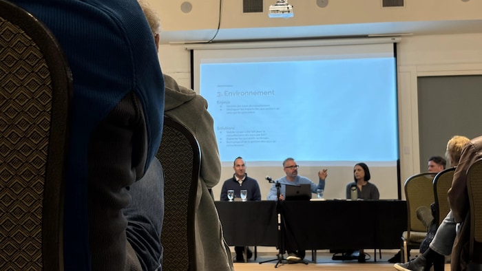 Une table avec 3 personnes au micro. Devant eux, des gens assis.
