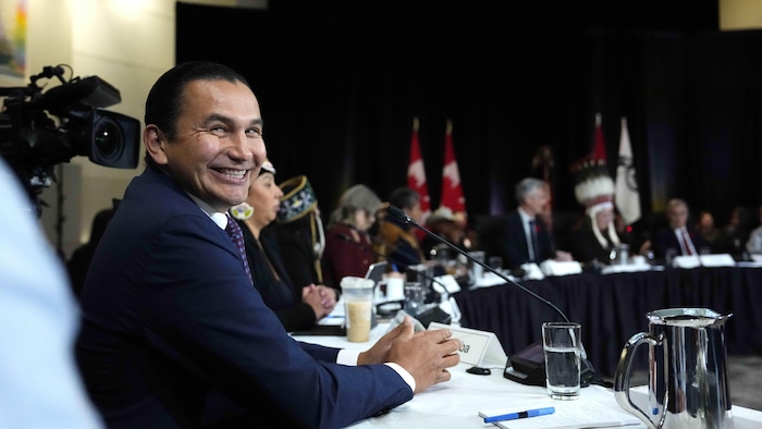 Wab Kinew souriant assis à une table.
