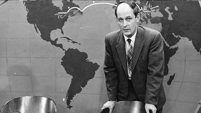 Debout devant une carte du monde, l'animateur René Lévesque est appuyé au dossier d'une chaise, une cigarette entre les
doigts.
