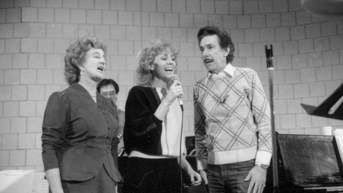 Renée Martel tient le micro et chante entourée de Noëlla Therrien et de Marcel Martel.