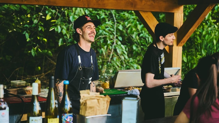 Un chef cuisinier sourit à des clients sous un gazebo extérieur.