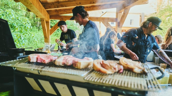 Trois chefs préparent des repas pendant que des pièces de viande cuisent sur une plaque de cuisson.