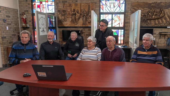 Un groupe de retraité est assis derrière une grande table avec un ordinateur, et regardent tous dans la même direction.