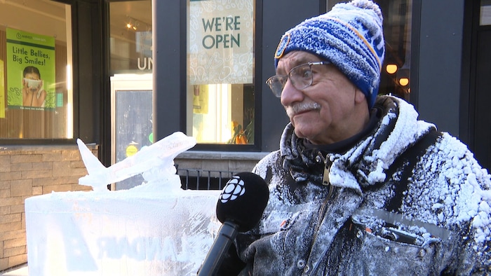 Un homme couvert de neige accorde une entrevue à côté d'un petit avion sculpté dans la glace.