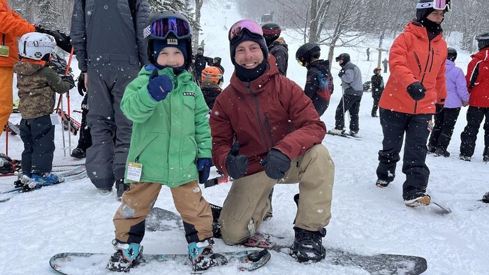 Richard Côté, un planchiste, est à genou à côté de son fils, aussi habillé en tenue de planche à neige. Ils sont en bas d'une piste au Mont-Comi. 