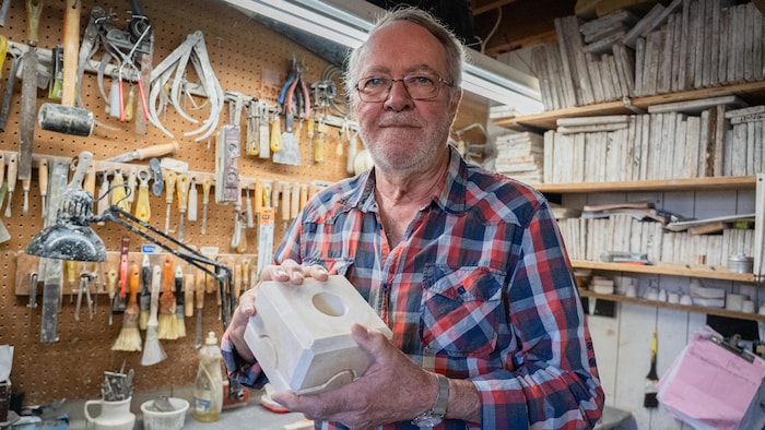 Un homme tient un moule de plâtre dans ses mains dans un atelier avec plusieurs outils accrochés derrière lui.