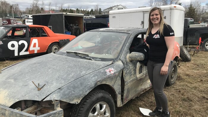Une femme se tient debout près d'une voiture toute sale et cabossée.