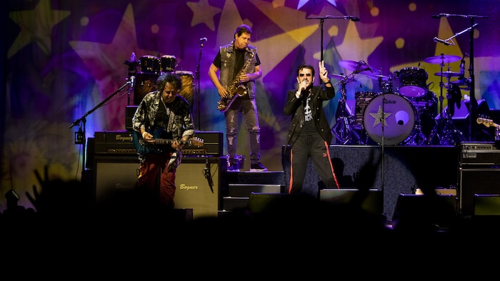 Ringo Starr chante sur scène. Il est accompagné d'un guitariste et d'un saxophoniste.