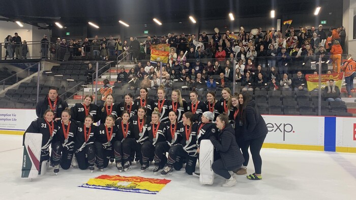 Les joueuses en uniforme posent pour une photo d'équipe sur la patinoire avec un drapeau du Nouveau-Brunswick devant elles.