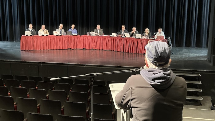 Un homme vu de dos parle au micro dans un amphithéâtre. Sur la scène devant lui, neuf personnes sont assis à une longue table et lui font face.