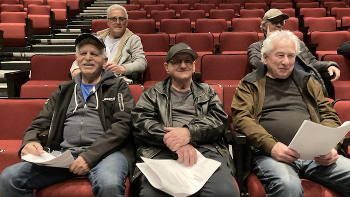 Cinq hommes vus de face sont assis dans un amphithéâtre. Ils ont des documents dans les mains.