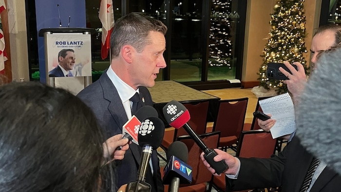 Rob Lantz de profil, entouré de journalistes, dans une salle décorée d'un sapin de Noël qui se reflète dans la fenêtre.