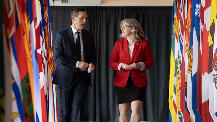 S'avançant vers la caméra, Rob Lantz et Susan Holt marchent entre deux rangées de grands drapeaux des provinces. Il parle et elle tourne la tête vers lui.