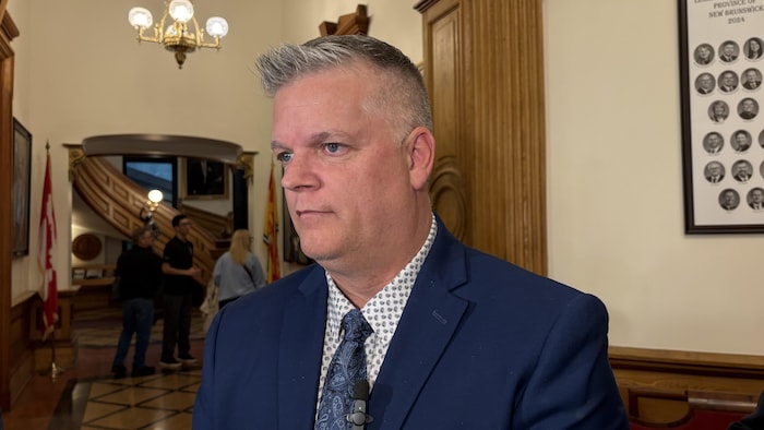 Une personne portant un complet bleu répond aux questions des journalistes dans la rodonde de l'Assemblée législative au Nouveau-Brunswick.