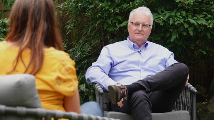 Robert Grant, en entrevue, assis sur une chaise dans son jardin, écoute la journaliste.