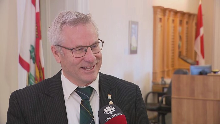 Le député est tourné vers la droite et parle, un micro pointé vers lui, dans un couloir de Province House.